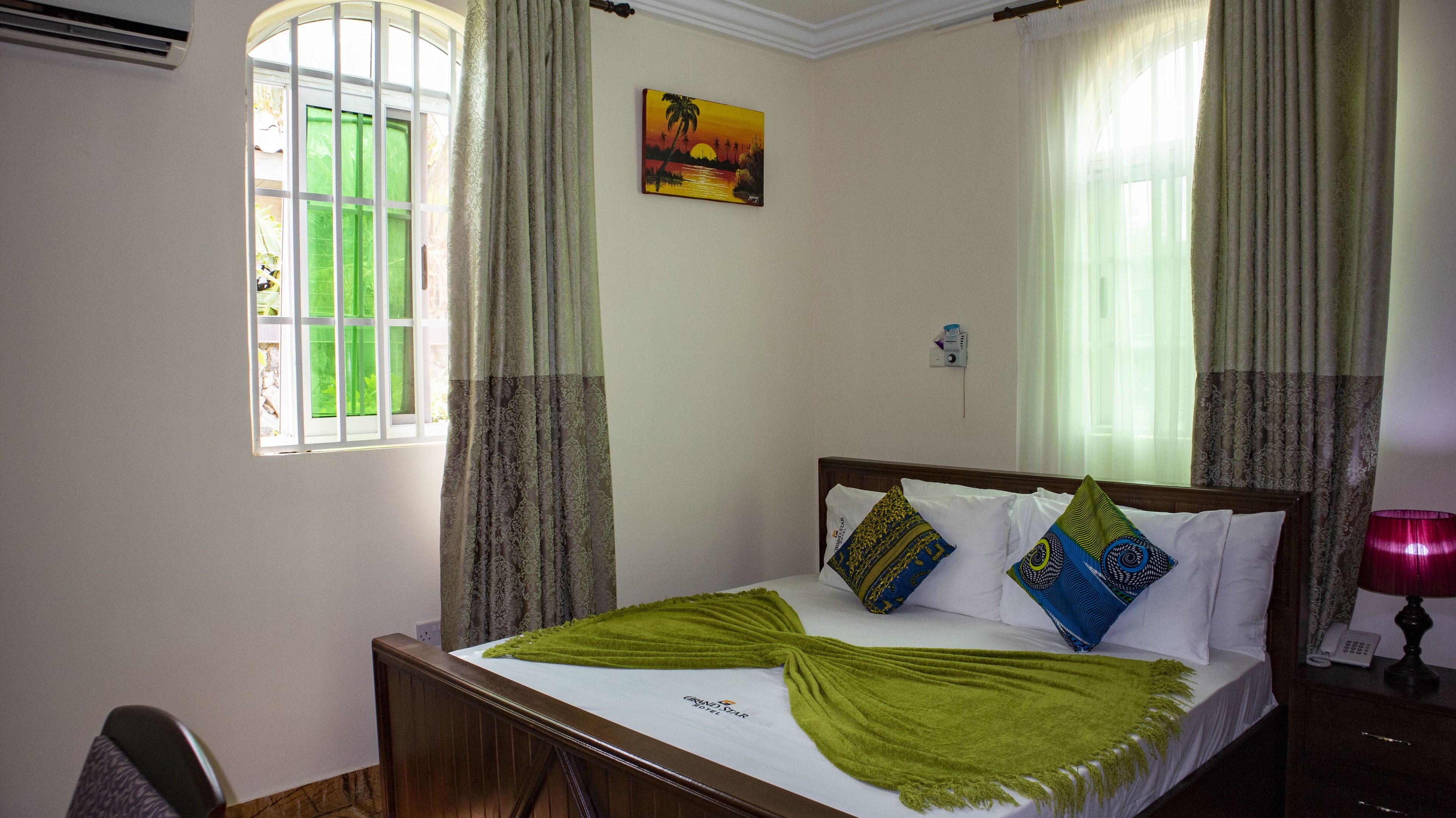 A hotel room with a bed, a painting, a window with curtains, and a bedside lamp.