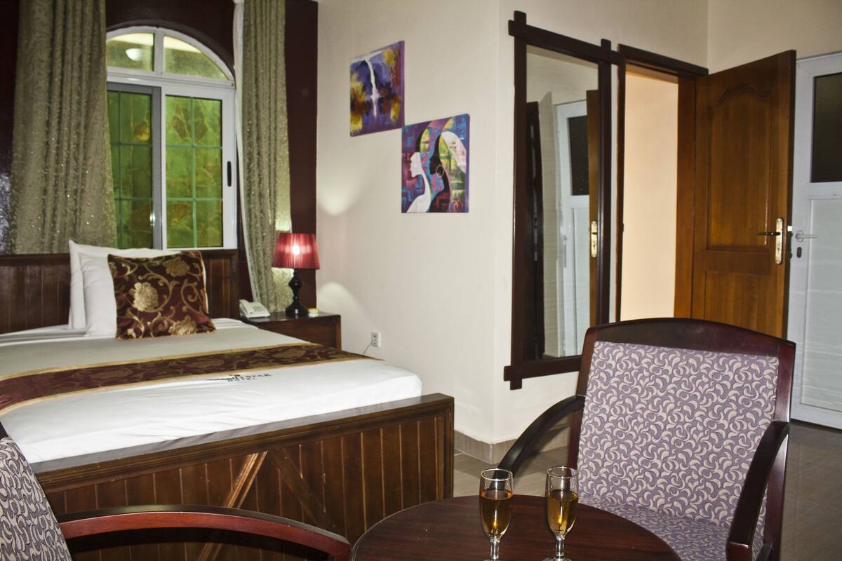 A hotel room with a bed, a chair, a table with two glasses of wine, and two paintings on the wall.