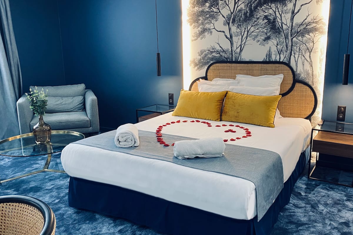 A modern hotel room with a large bed, a side table, a chair, and a wall-mounted painting.