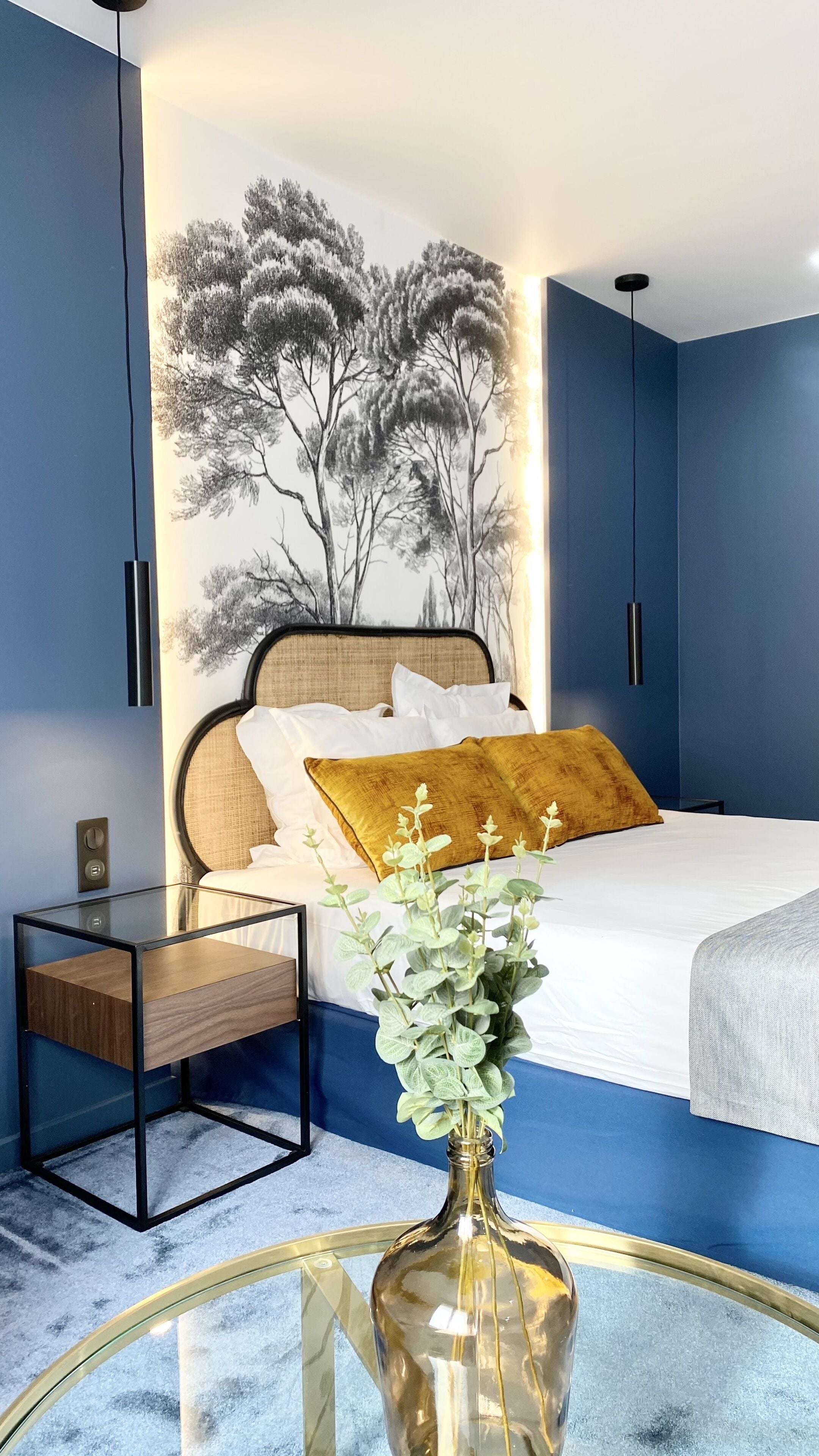 Quarto com cama, mesa de cabeceira, vaso com flores e parede com painel de árvore.