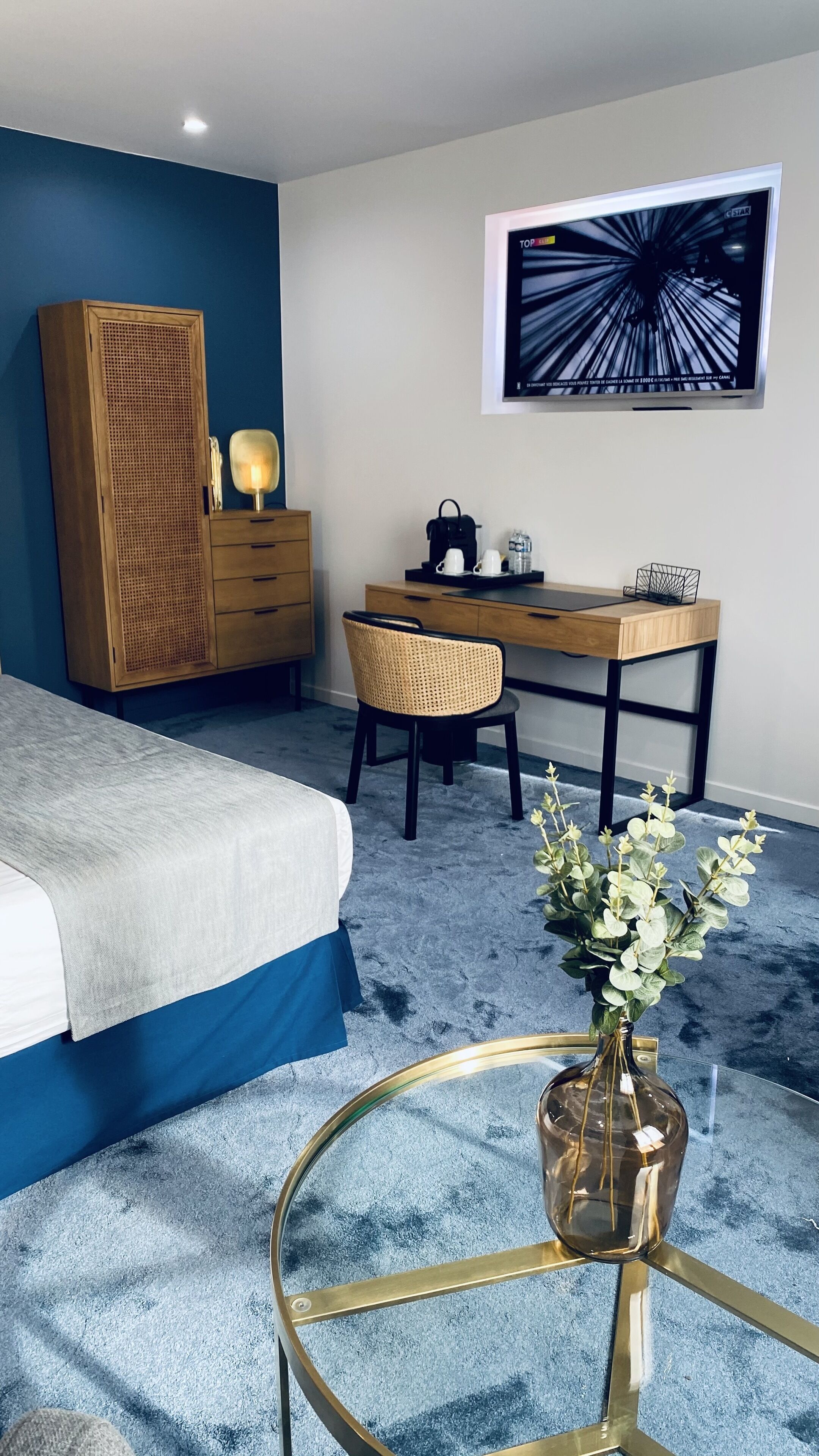 Quarto com cama, escrivaninha, cadeira, guarda-roupa, mesa de centro e televisão fixada na parede.