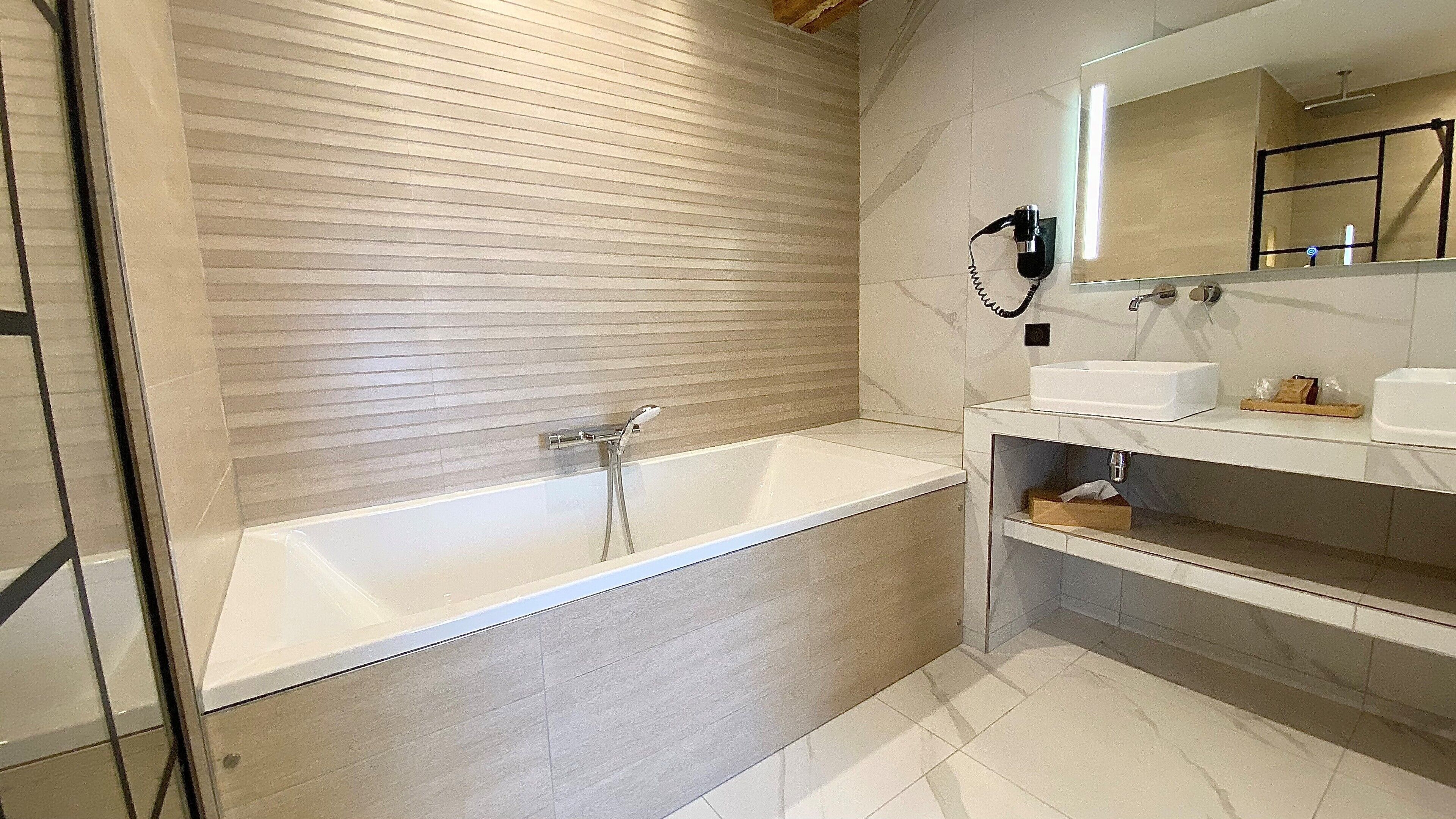 A modern bathroom with a large bathtub, a vanity with a mirror, and a hairdryer mounted on the wall.