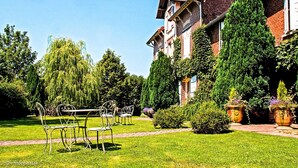 Un jardín con una mesa y sillas de metal, un edificio de ladrillo y árboles altos.