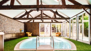 Una piscina interior con pared de ladrillo, plataforma de madera y puertas de cristal que dan al exterior.