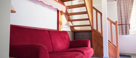 Un canapé rouge dans une pièce avec un escalier en bois et une fenêtre ornée d’un rideau à carreaux.