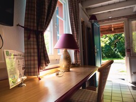 Una mesa de madera con una lámpara, un letrero y una silla en una habitación con cortinas a cuadros y vistas a la vegetación exterior.