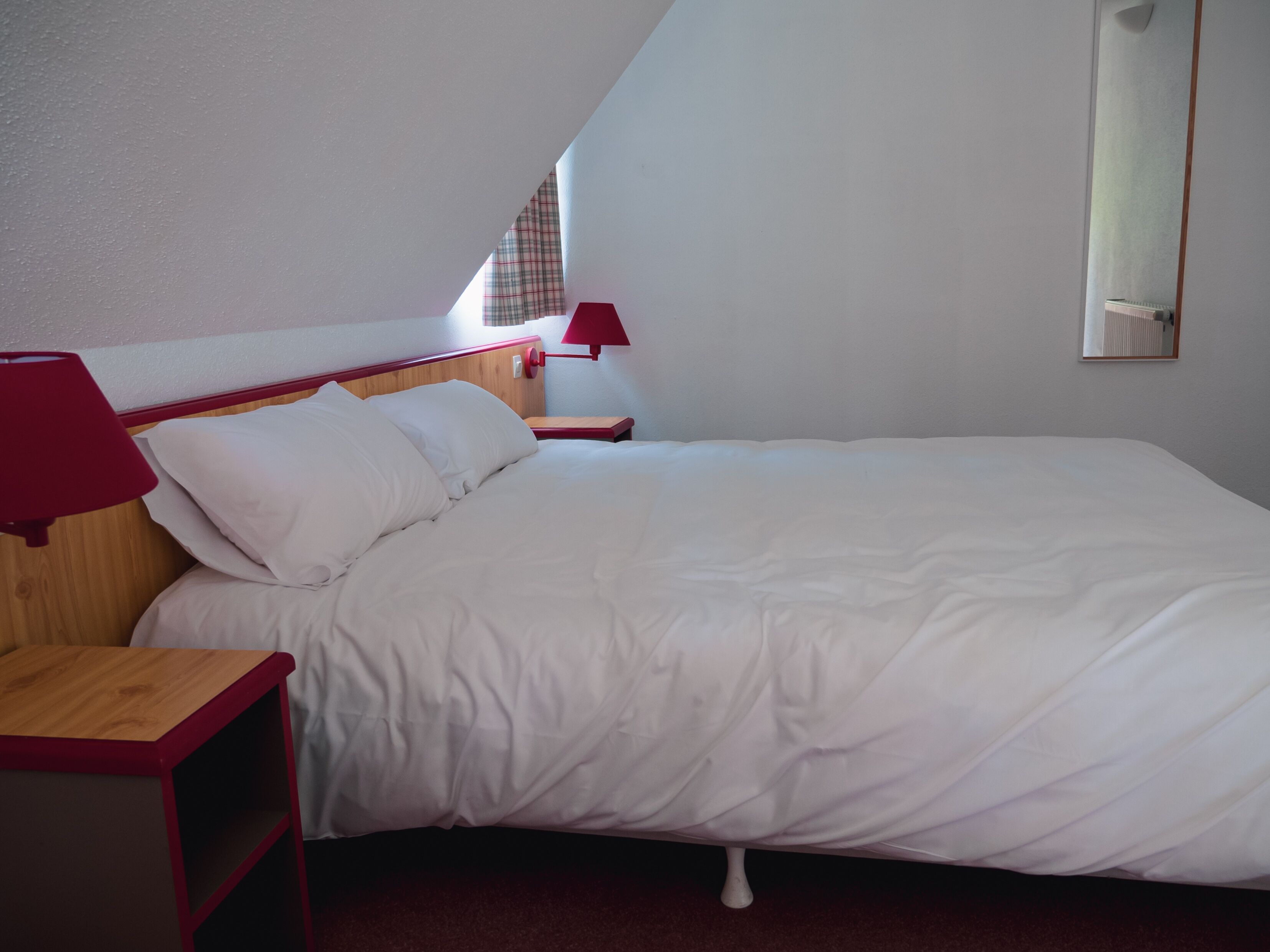 Een slaapkamer met een enkel bed, een nachtkastje, een rode lamp en een raam met een geruit gordijn.