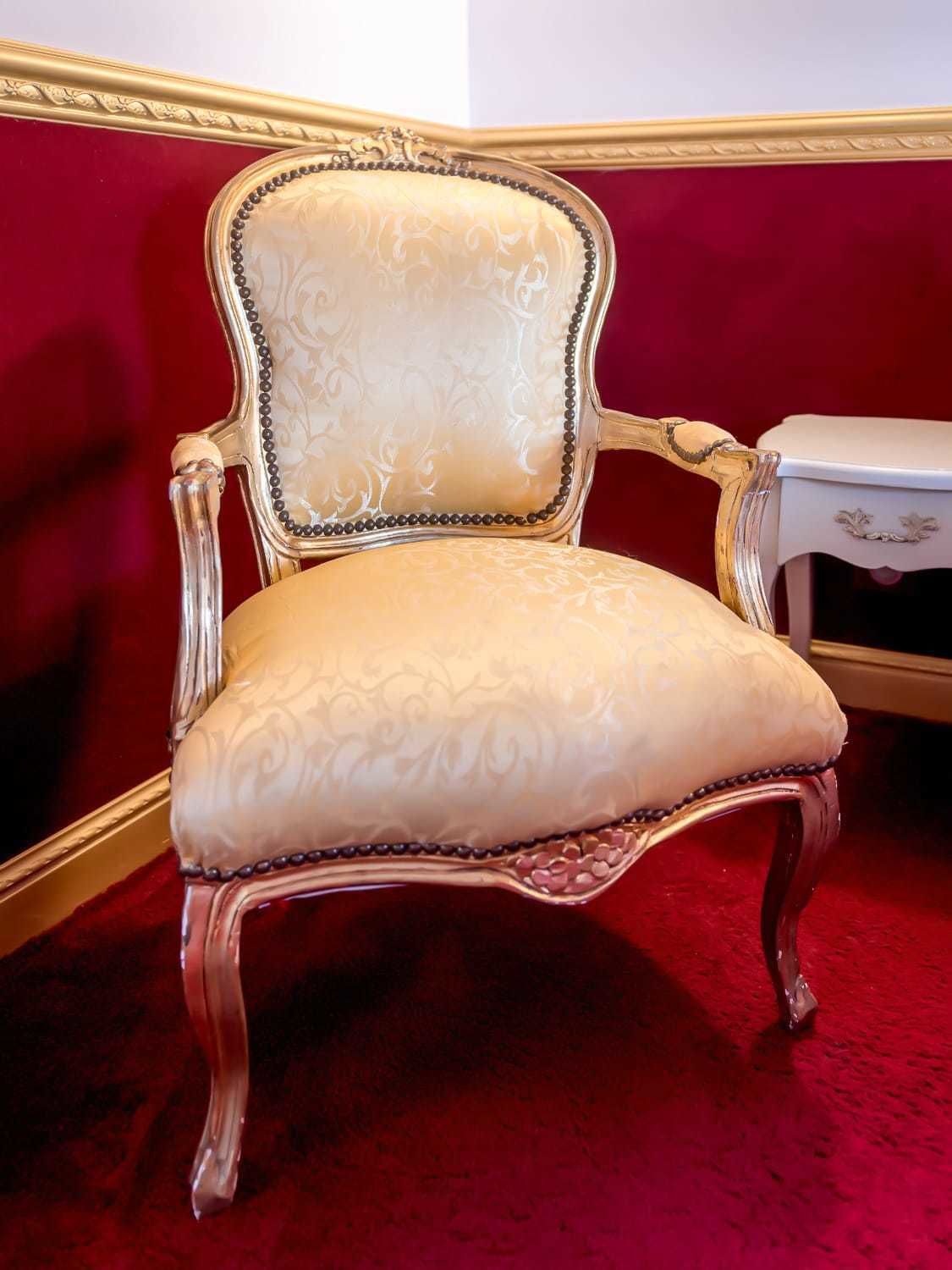 A luxurious armchair with intricate gold detailing and a plush, patterned seat, positioned against a red carpeted floor and a white side table.