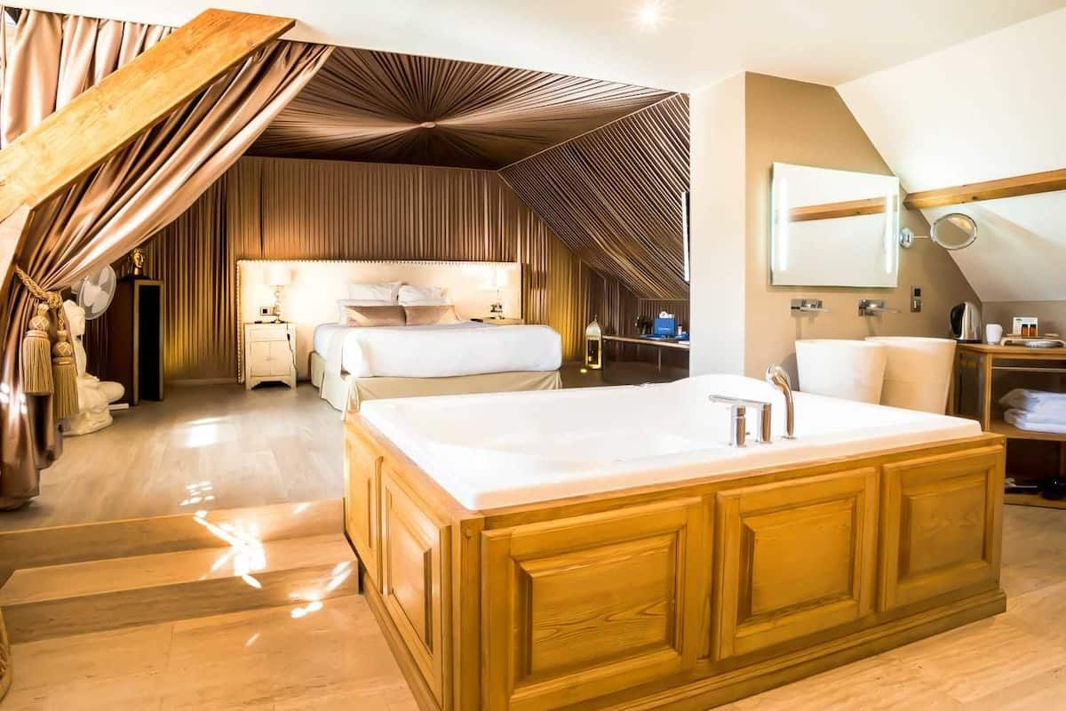 A modern hotel room with a large wooden vanity, a separate bedroom with a bed and bedside tables, and a bathroom with a large mirror and sink.