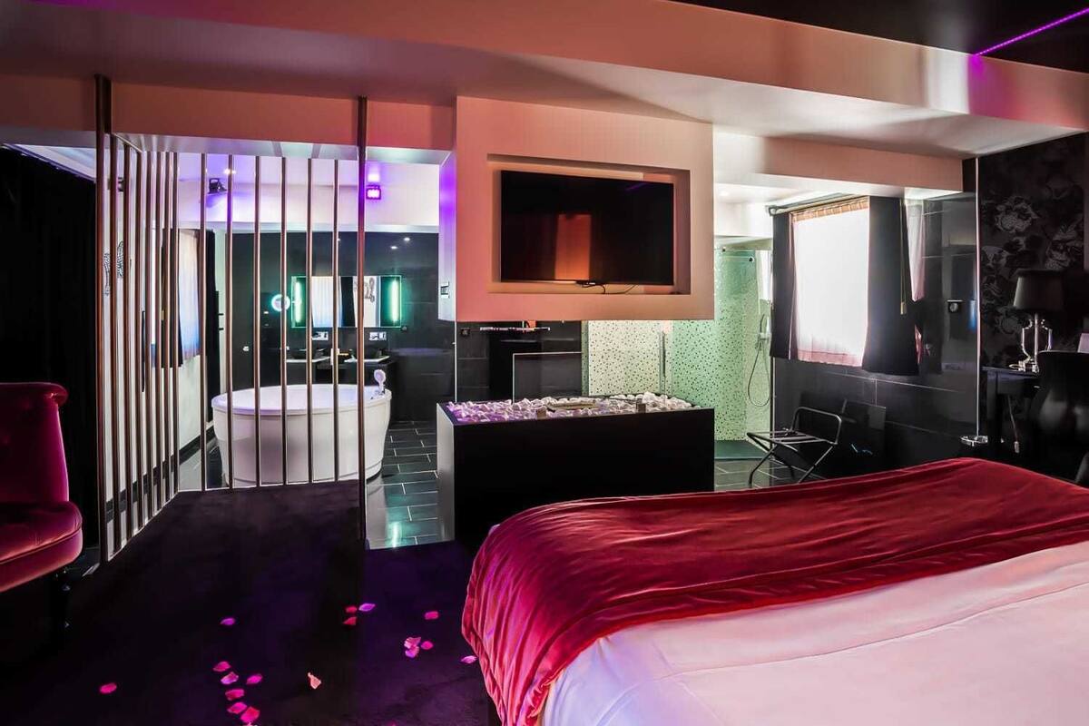 A modern hotel room with a large bed, a TV, a desk, and a bathroom visible through a partition.