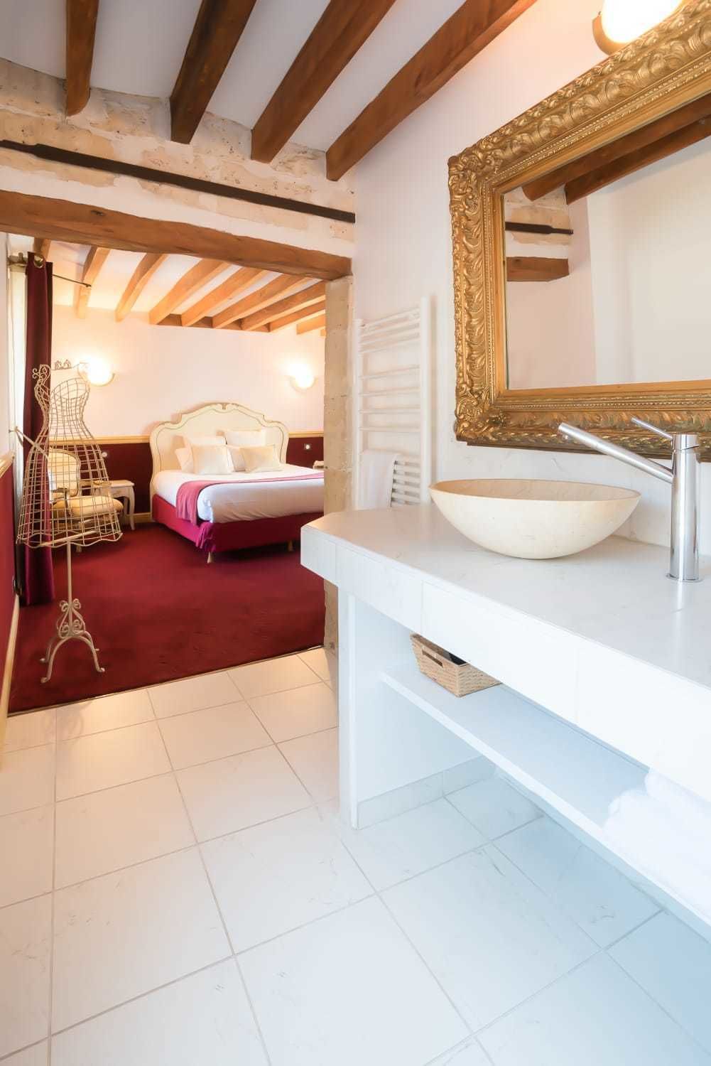 Habitación de hotel con una cama grande, un tocador blanco con lavabo, un espejo con marco dorado y vigas de madera a la vista en el techo.