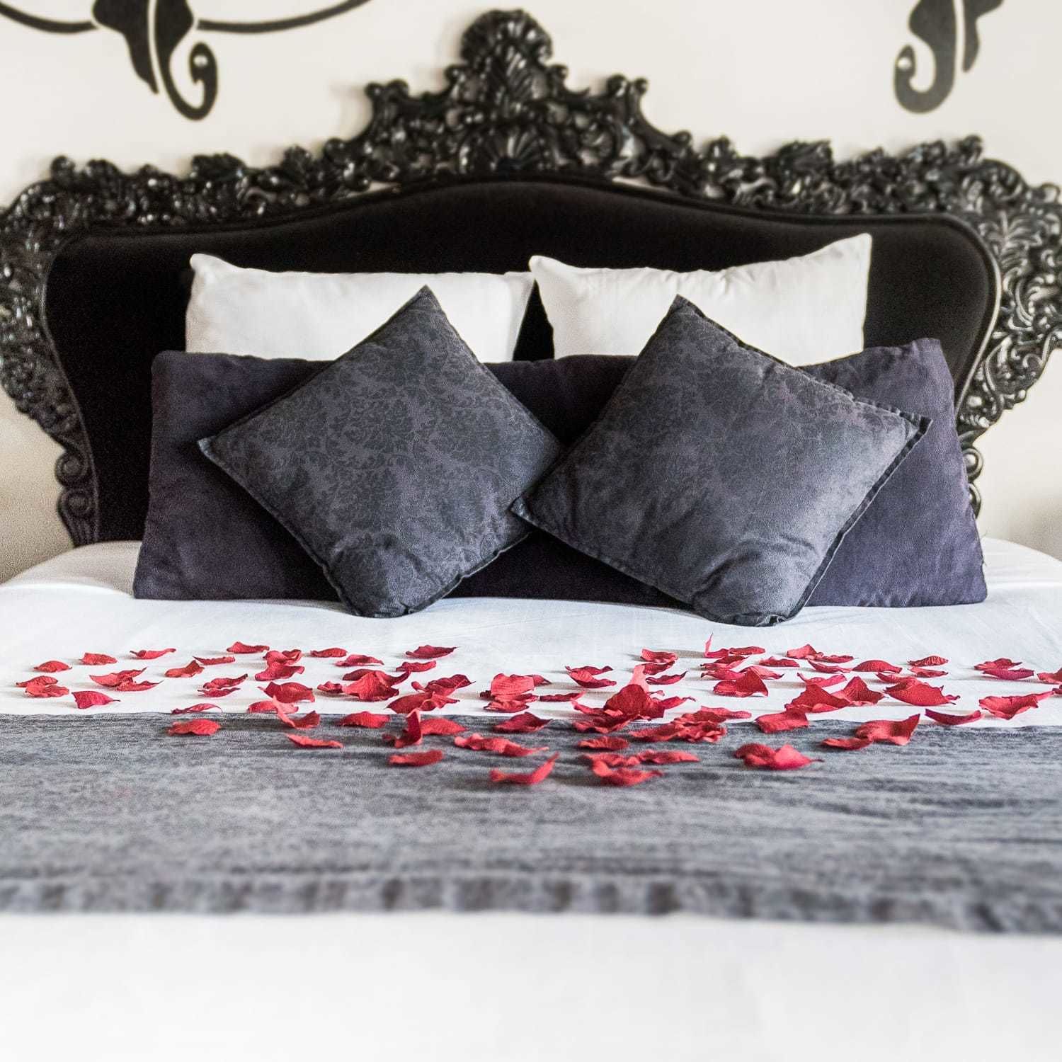 Una cama con cabecera negra, dos almohadas oscuras y pétalos de rosa roja esparcidos sobre un cubrecolcha blanco.
