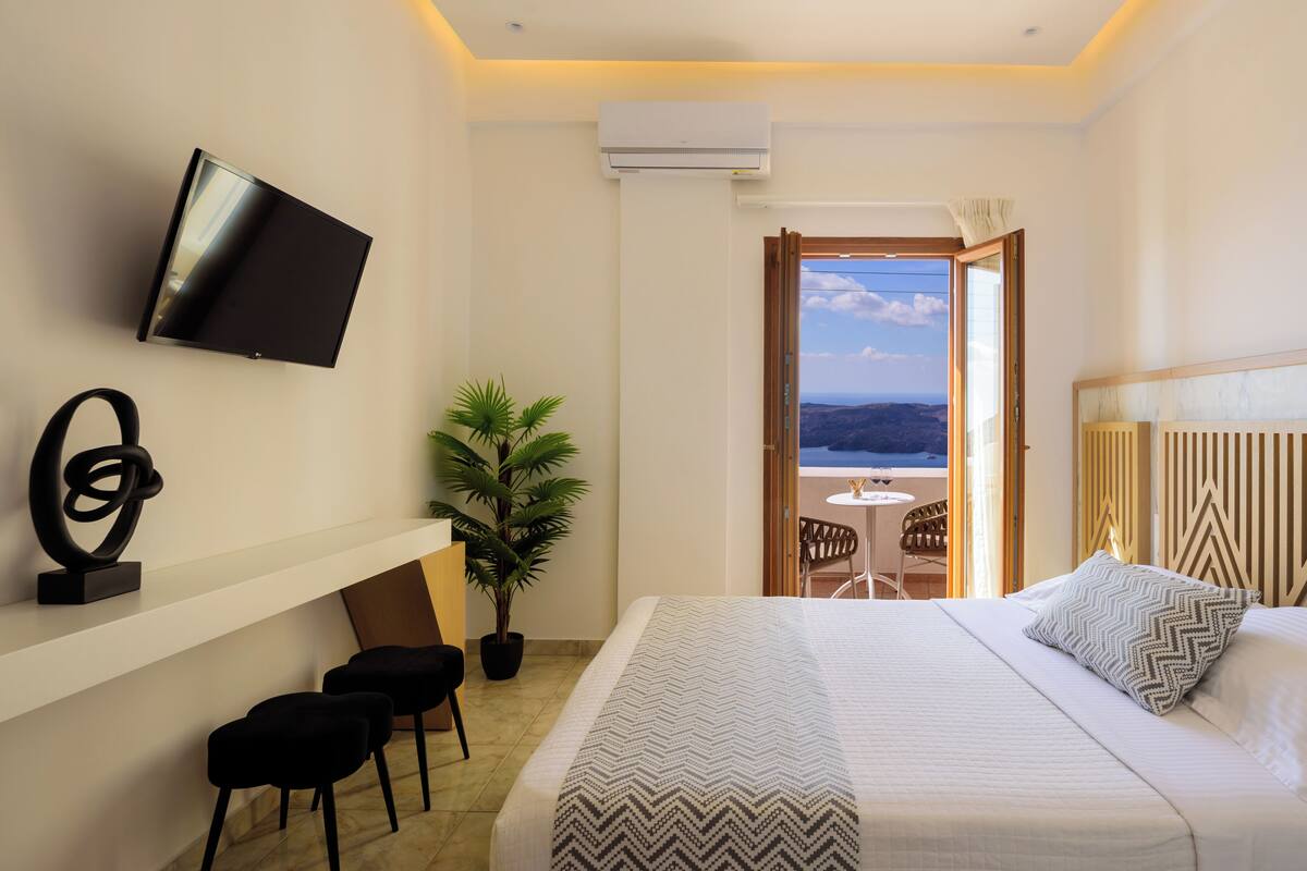 A hotel room with a bed, a TV mounted on the wall, a desk with chairs, a potted plant, and a balcony with a view of the landscape.