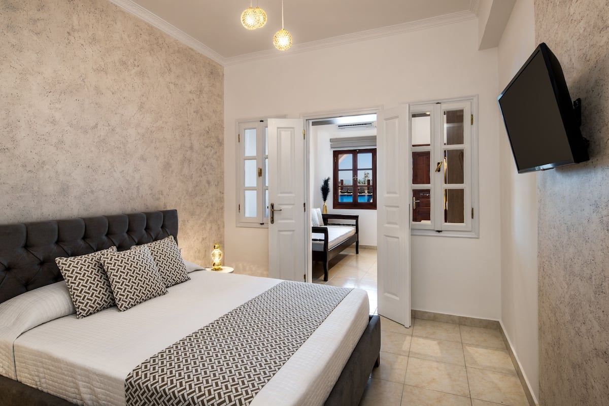 A bedroom with a large bed, a TV mounted on the wall, and a view of a pool through an open door.
