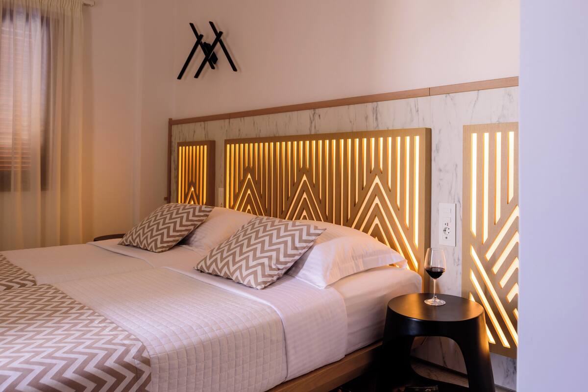 A neatly made bed with patterned pillows, a bedside table with a glass of wine, and a wall with geometric light panels.