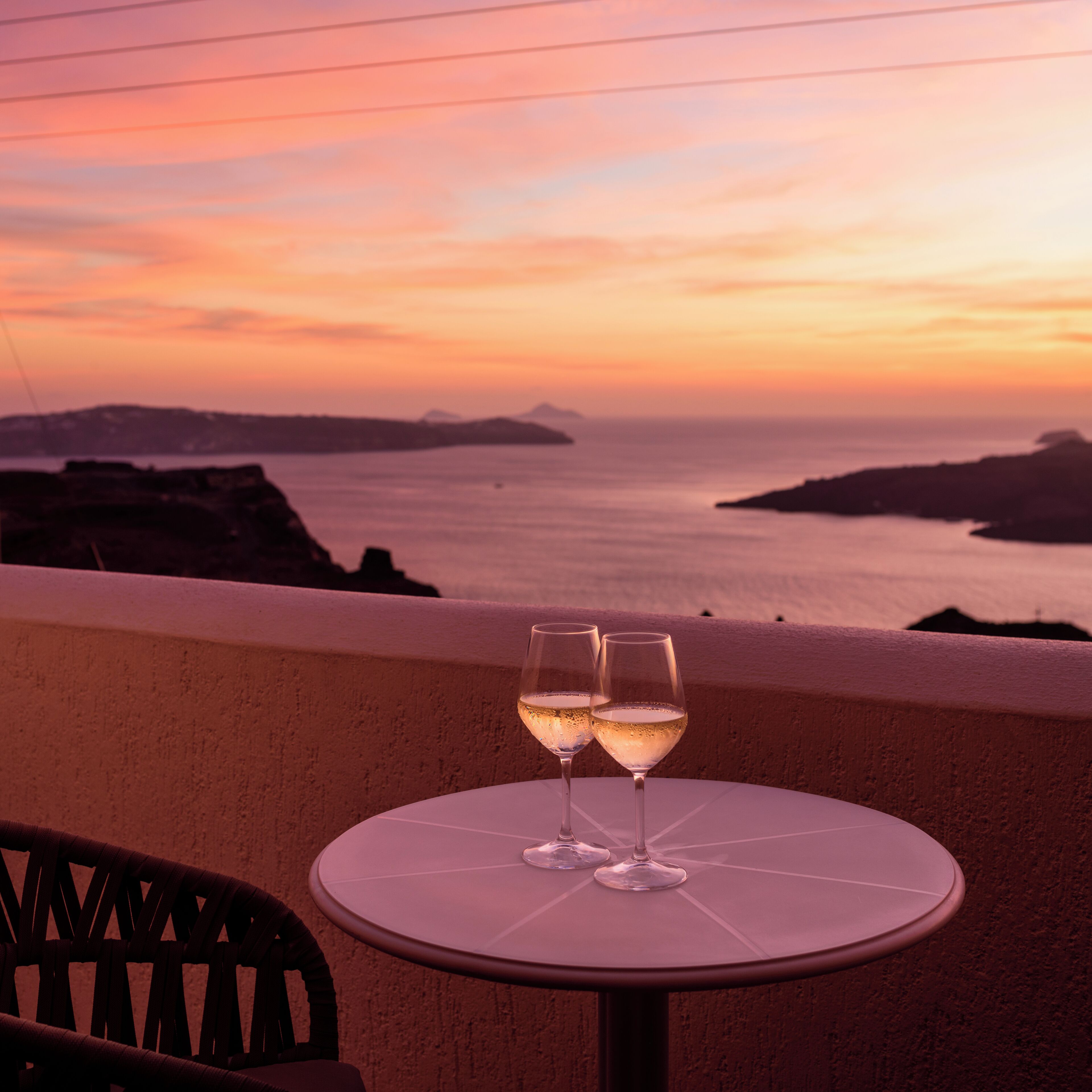 Un tavolo con due bicchieri di vino su un balcone affacciato su un tramonto sul mare.
