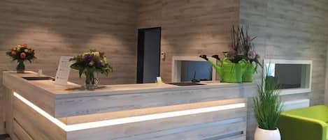 Egy fa borítású recepciós pult, egy zöld szék és virágos növények cserépben.