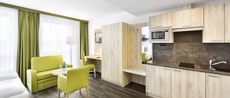 A modern hotel room with a green sofa, a small dining table, a kitchenette with a microwave, and a large window with green curtains.
