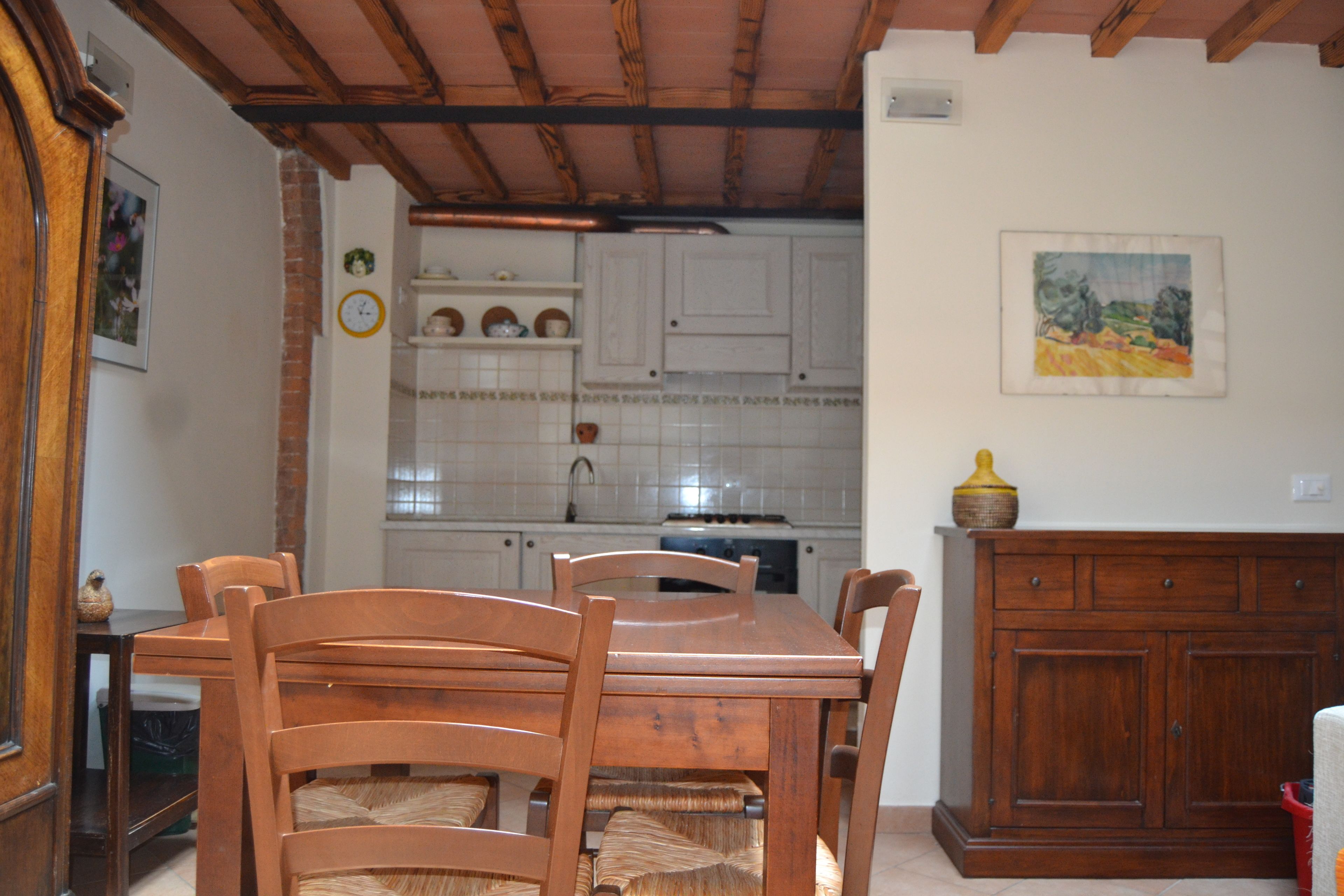 Un coin repas avec une table et des chaises en bois, une cuisine à l’arrière-plan et un tableau accroché au mur.