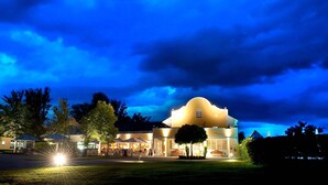A large, yellow building with a curved roof, surrounded by trees and a well-lit outdoor area with umbrellas. - Golfhotel Gutshof Penning (Rotthalmuenster)