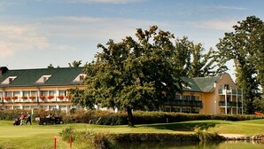 A golf course with a pond, a large tree, and a hotel with multiple balconies. - Golfhotel Gutshof Penning (Rotthalmuenster)
