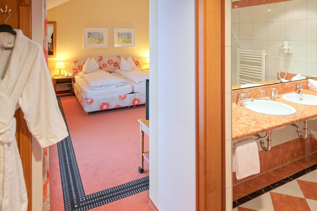 A hotel room with a bed, two bedside tables, a nightstand, a bathroom with a sink and mirror, and a hallway with a robe hanging.
