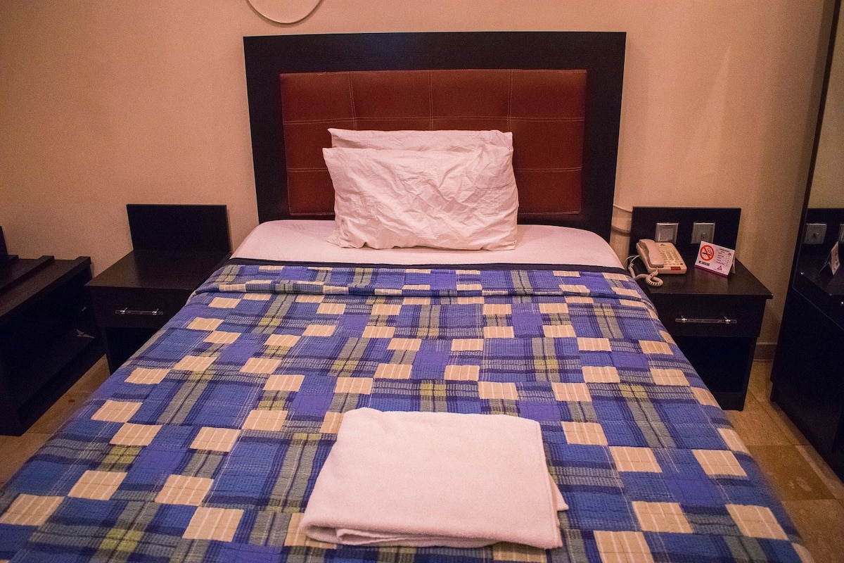 Quarto de hotel com uma cama grande, cabeceira, mesa de cabeceira com telefone e relógio fixado na parede.