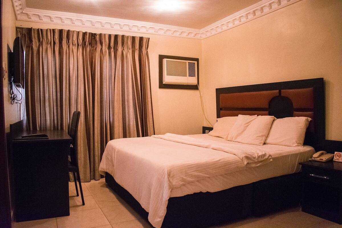 Quarto de hotel com uma cama, escrivaninha, cadeira, ar condicionado e mesa de cabeceira com telefone.