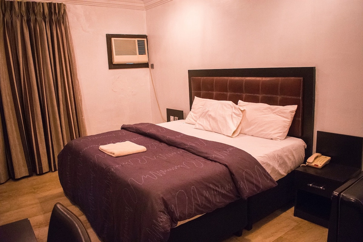 Quarto de hotel com uma cama grande, uma mesa de cabeceira, uma cadeira e uma cortina.