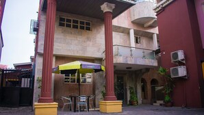 A building with a red and beige facade, featuring a balcony, a covered outdoor seating area with chairs and an umbrella, and a potted plant. - Maidaville Hotel and Suites (Lagos)