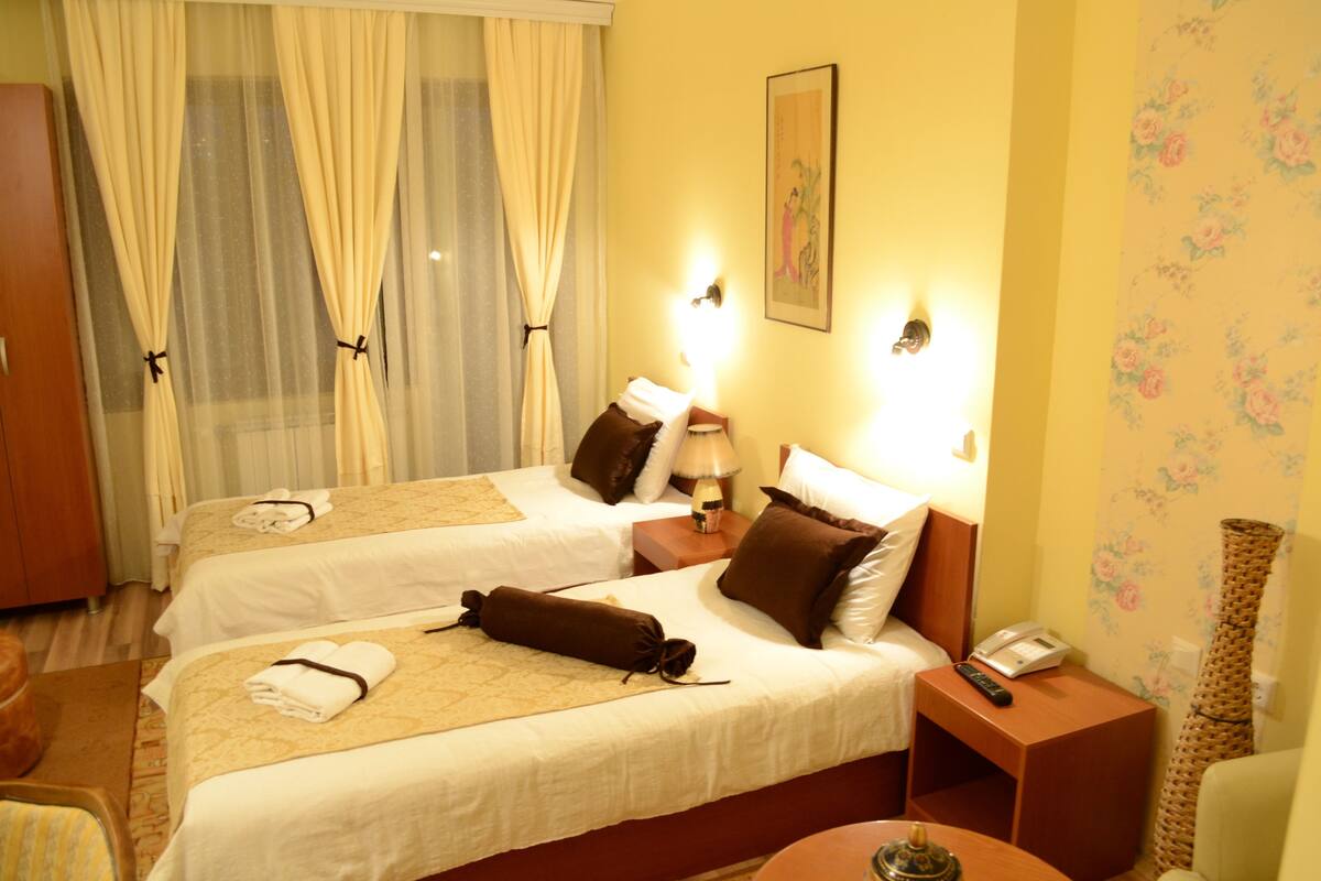 A hotel room with two beds, a bedside table with a lamp, a telephone, and a decorative vase.
