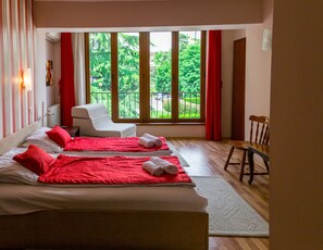 A hotel room with two beds, a chair, a wooden table, and a large window with a view of greenery. - Hotel Pine (Skopje)
