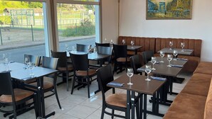 Um restaurante com várias mesas e cabines, grandes janelas que oferecem vista para o exterior, e luminárias pendentes.