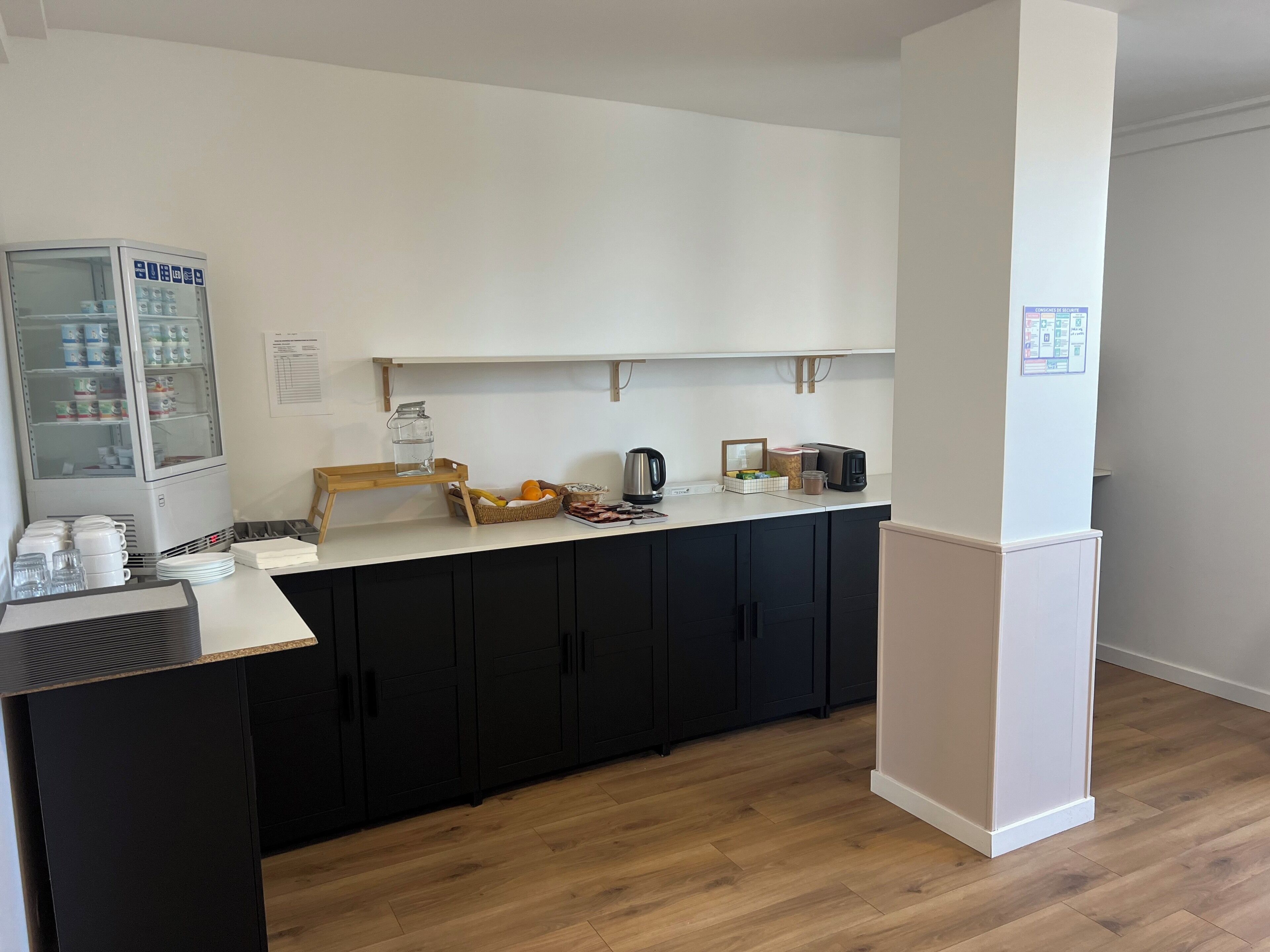 Una kitchenette equipada con refrigerador, microondas y cafetera. Encima del mostrador hay un estante de madera con varios objetos.