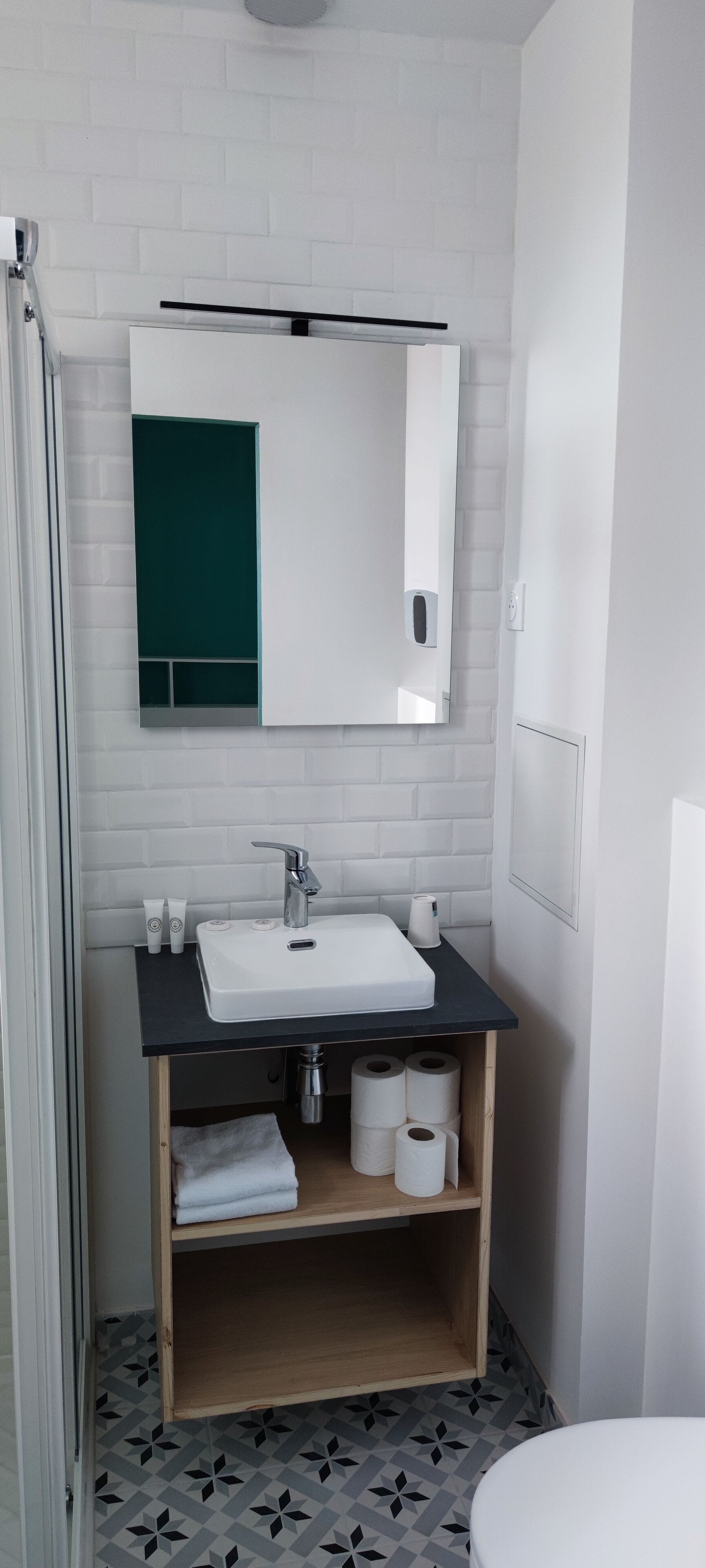 A modern bathroom with a white sink, dark countertop, and a mirror above it. There are two shelves below the sink, one holding a towel and the other with toilet paper.