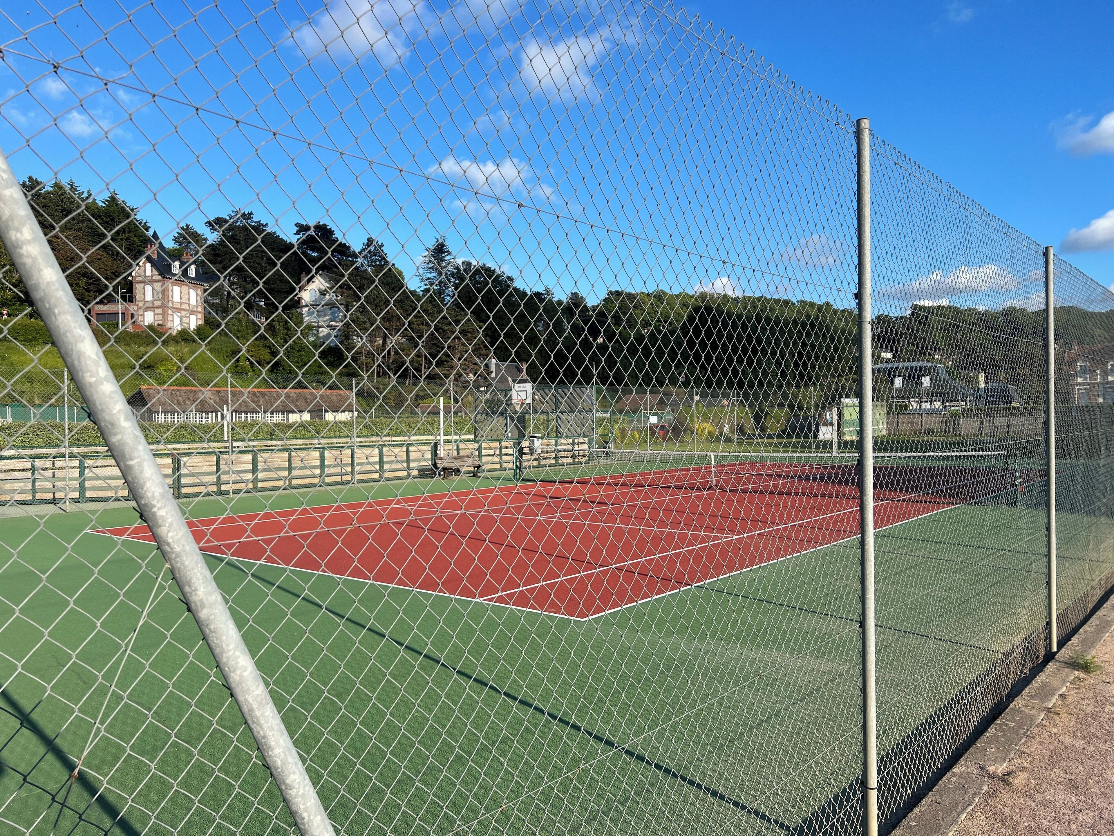 Un terrain de tennis délimité par une clôture en treillis métallique, doté d’une surface verte et d’une zone rouge, et entouré d’arbres et de bâtiments.