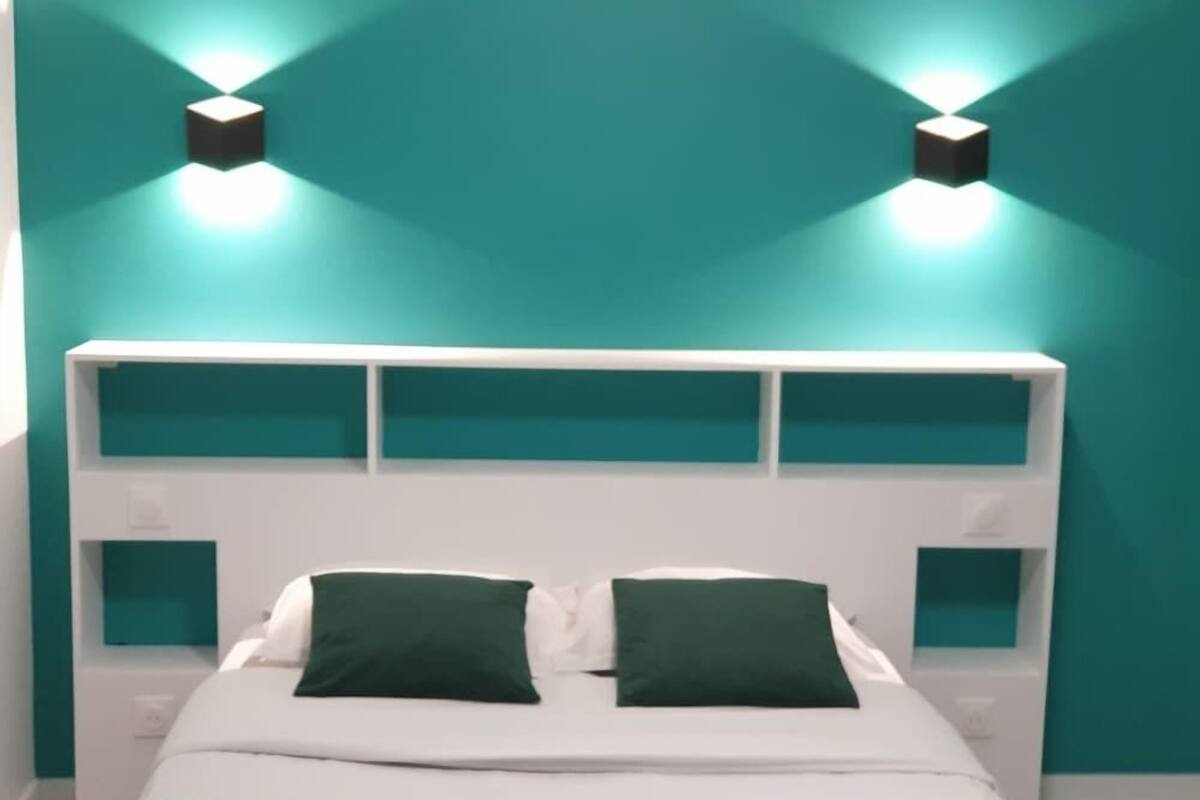 Un lit avec une tête de lit blanche, deux oreillers verts et un mur de couleur turquoise agrémenté de deux appliques murales.