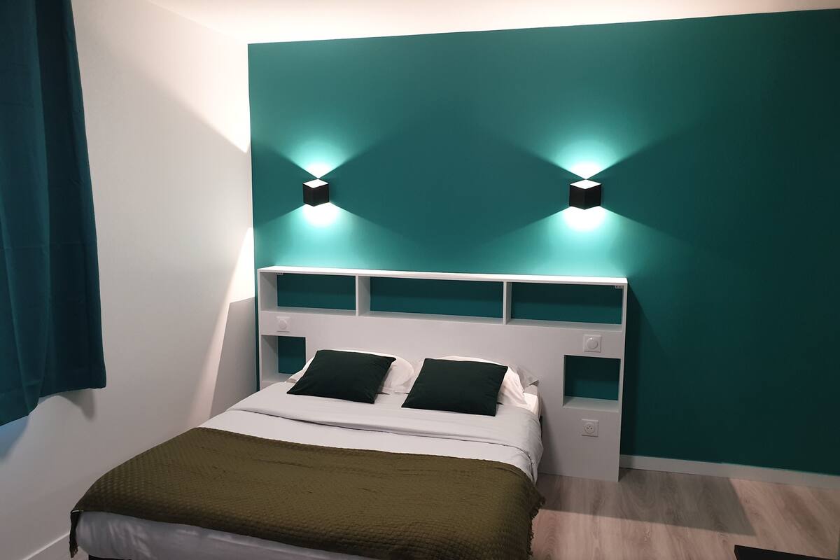 Un lit bien fait, agrémenté d’un plaid vert, de draps blancs et d’une tête de lit blanche dotée d’étagères.