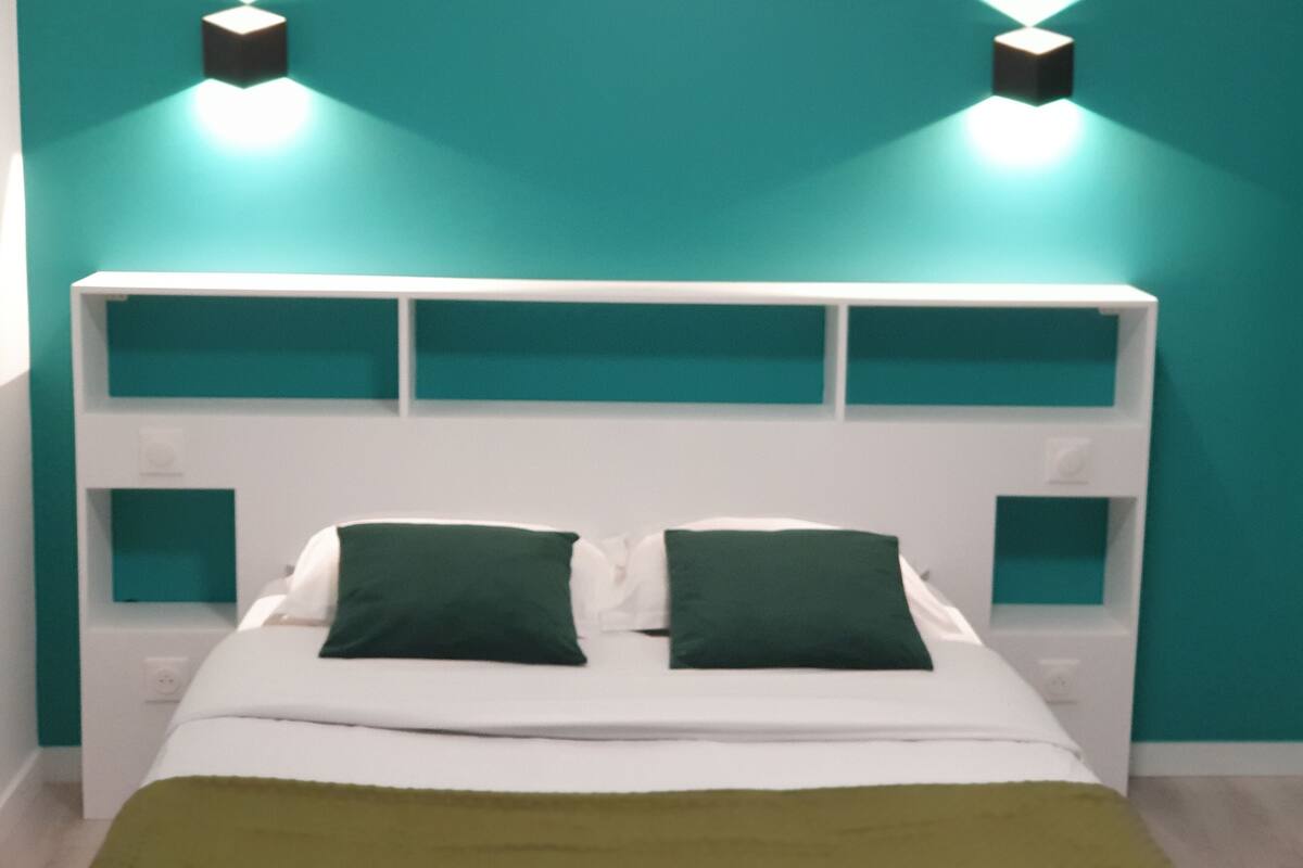 Un lit avec une tête de lit blanche et des murs de couleur turquoise, deux coussins verts et deux appliques murales.