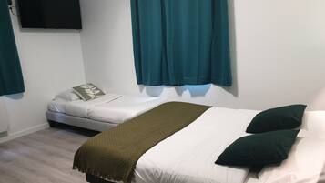 Habitación con dos camas, una con una manta verde y la otra con ropa de cama blanca. Hay un televisor de pantalla plana montado en la pared y cortinas color turquesa en las ventanas.