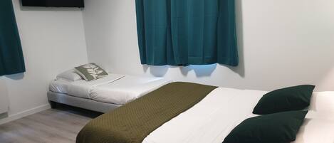 Habitación con dos camas, una con una manta verde y la otra con ropa de cama blanca. Hay un televisor de pantalla plana montado en la pared y cortinas color turquesa en las ventanas.