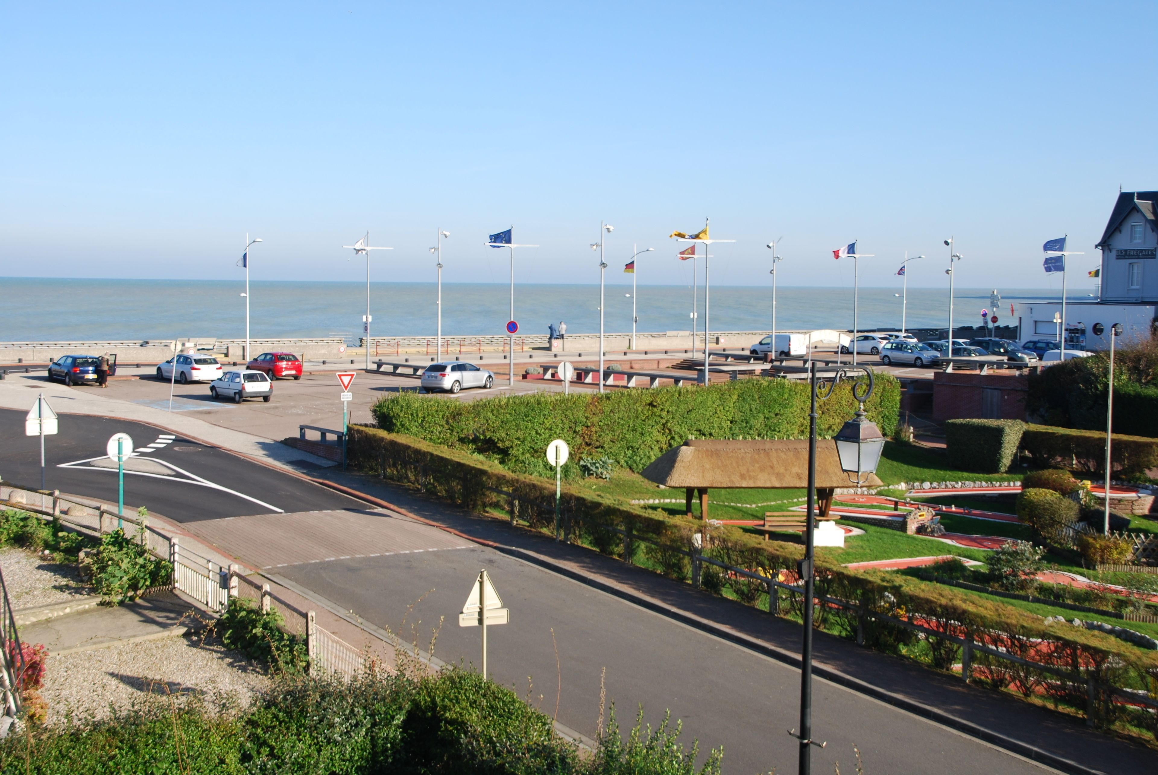 A coastal road with parked cars, a pedestrian crossing, and a thatched-roof pavilion.
