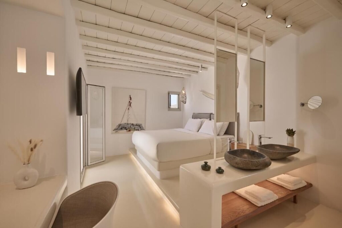 A modern bedroom with a large bed, bedside tables, and a bathroom with a large mirror and two bowls.
