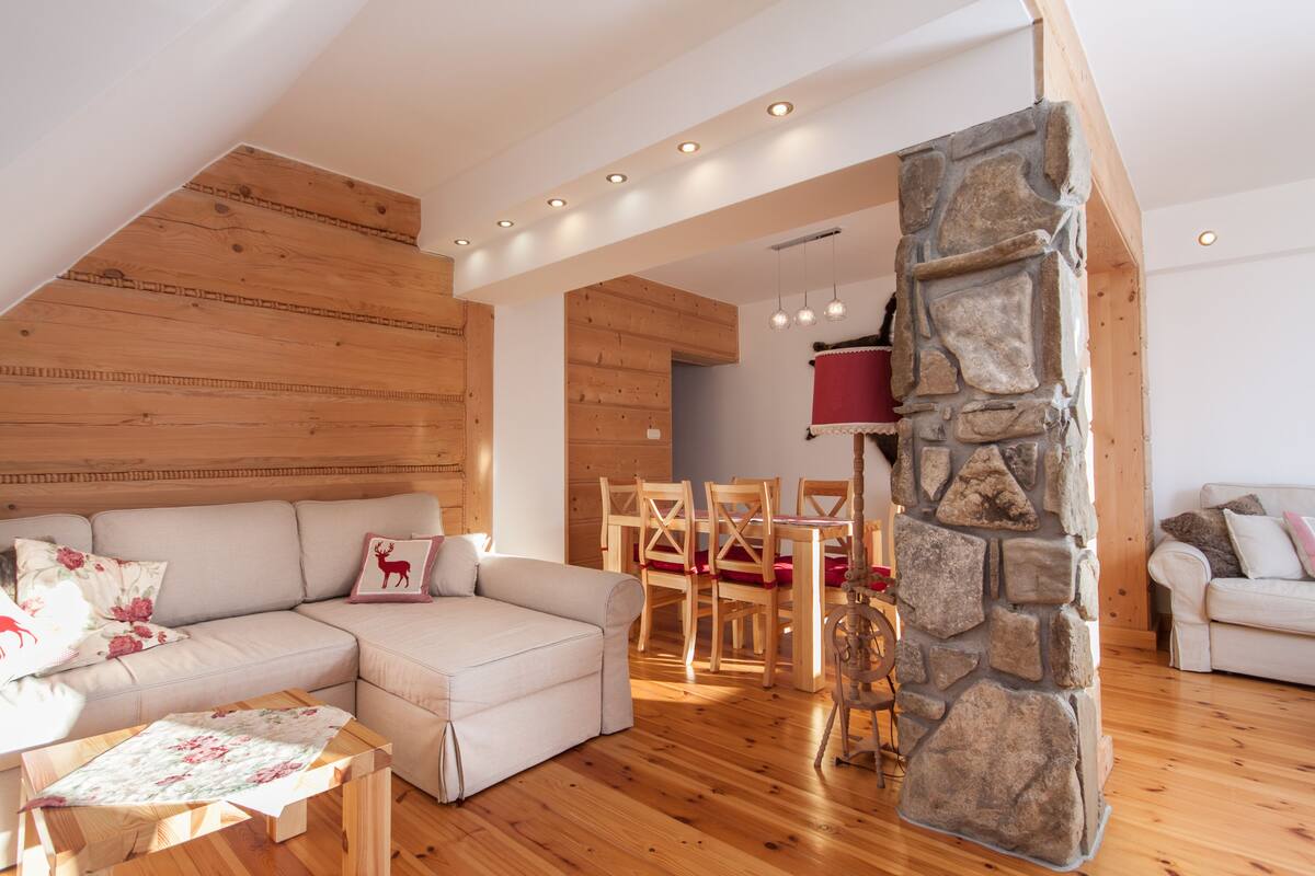 A cozy living room with a wooden ceiling, a stone fireplace, a sofa, and a dining area with wooden furniture.