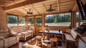 Ein gemütliches Blockhäuschen-Interieur mit Ledersofa, Holzesstisch und großen Fenstern mit Blick auf den Wald.