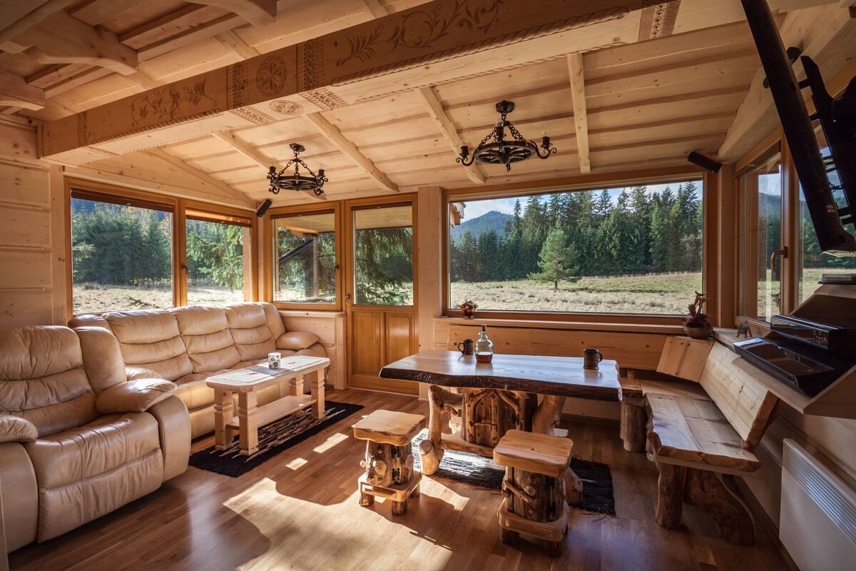 A cozy cabin interior with a leather sofa, wooden dining table, and large windows offering a view of the forest.