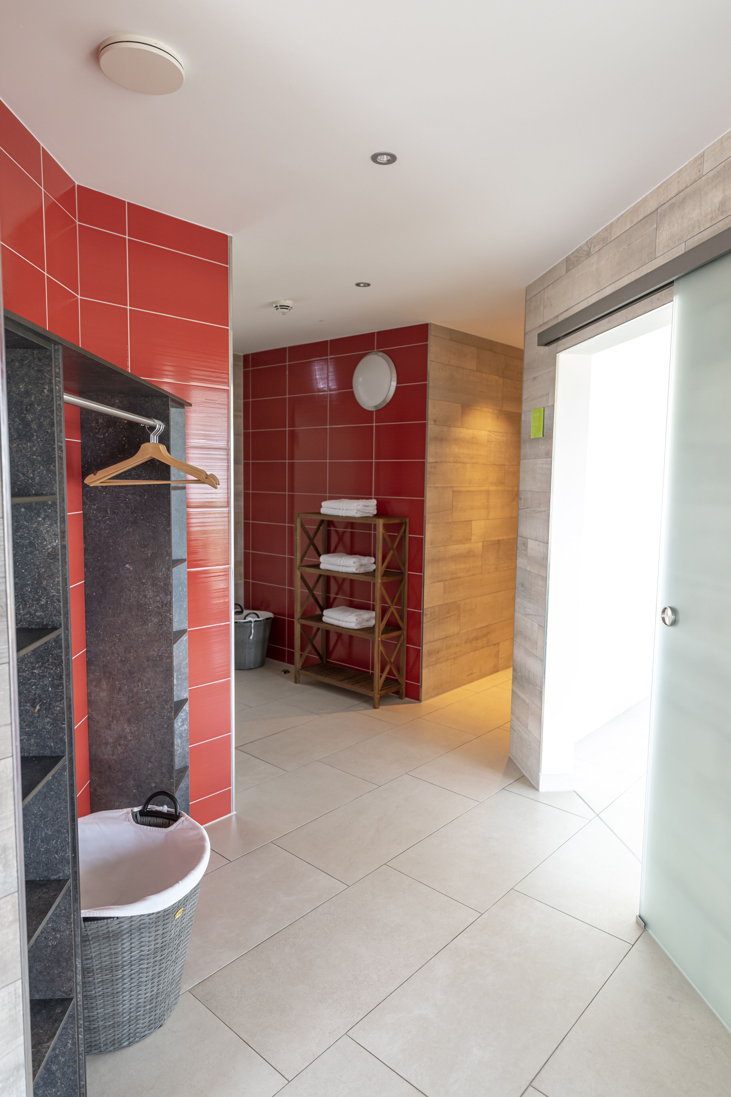 A bathroom with a red tiled wall, a wooden shelf with towels, a hanging rack, and a trash bin.