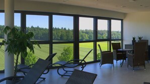 A room with large windows offering a view of a green landscape. There are wicker chairs and a small table with two potted plants. - Hotel Campo (Renningen)