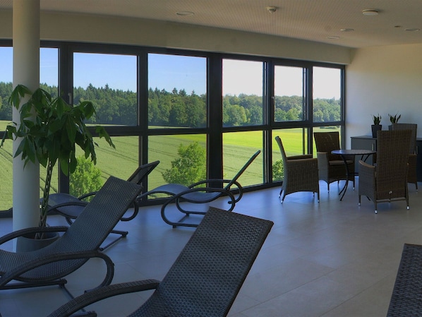 Habitación con ventanas amplias que ofrecen vistas a un paisaje verde. Hay sillas de mimbre y una mesita baja con dos plantas en macetas.