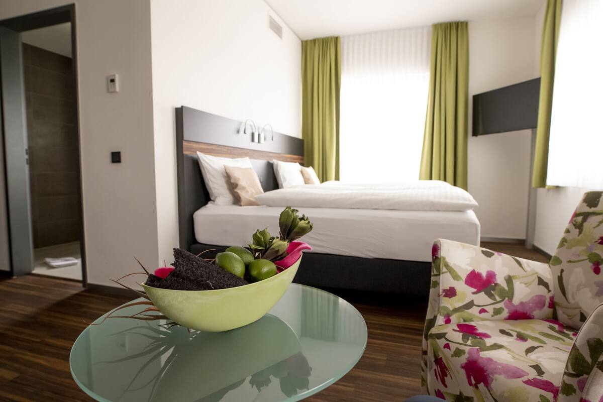 A hotel room with a bed, a glass table with a bowl of plants, a floral armchair, and a television on the wall.
